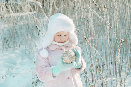 Little cute smiling girl in pink coat standing in the winter magic forest and holding Christmas or New Year decoration ball in her handsの写真素材