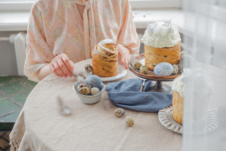 Beatiful woman hands with easter cakes on table with quail eggs and feathers, painted eggs, over windowの写真素材