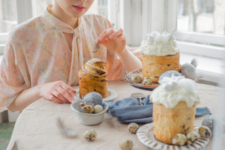 Beatiful woman hands with easter cakes on table with quail eggs and feathers, painted eggs, over windowの写真素材