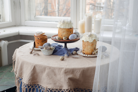 Easter cakes on table with quail eggs and feathers, painted eggs over window, rustic roomの写真素材