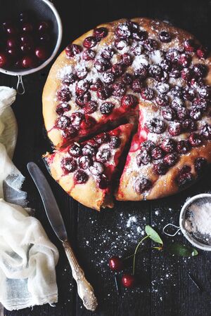 Dark and moody photo of Focaccia bread with cherries with a piece cut out on the black wooden background taken using natural lightの写真素材