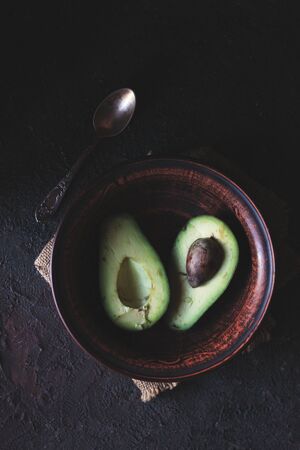 Green Avocado Halves in a Brown Clay Bowl With a Spoon Lying Around on a Black Backgroundの写真素材