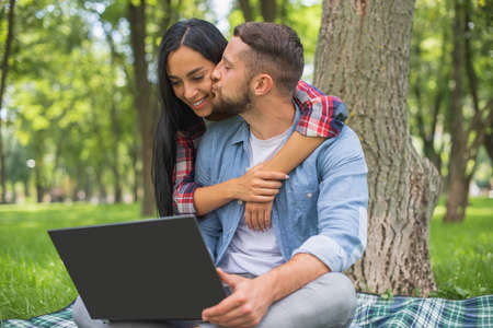 Love couple spends time in the park with a laptop. Kiss on cheek. Happy couple and joint activities. Joint hobby of a girl and a guy.の写真素材