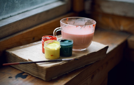 Still life of gouache paints, brushes, paint dilution cup and a book that teaches drawing. All items are on the old wooden windowsill.の写真素材