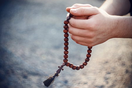 Man holding a wooden rosary and praying. Hands with rosary illuminated by sunlight.の写真素材