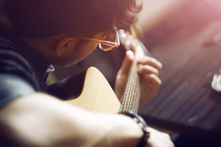 A curly-haired inspired guy in a shirt and pink glasses plays a melody on an acoustic guitar,...