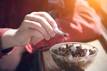 The man in the red plaid shirt sitting by the window, from which shines the sunlight, leaning arm on the window sill and smokes a cigarette, flicking the ashes into a dirty ashtray filled with cigarette butts.の写真素材
