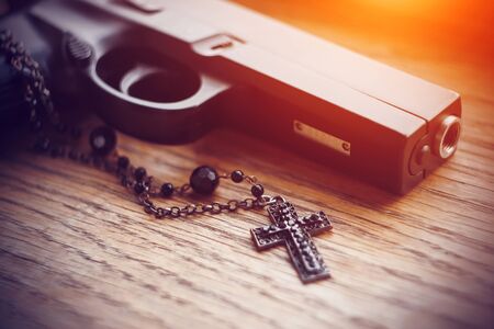 On the old wooden surface lies a black gun and a black cross on a chain with black beads, illuminated by a bright aggressive light.の写真素材