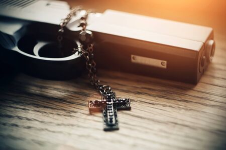 On a wooden table lies a gun lit by sunlight and black carved Holy cross with black beads on the...