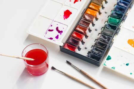 Watercolor palette with a lot of shades, a few fluffy brushes and a glass Cup of water for washing brushes, already stained red.の写真素材