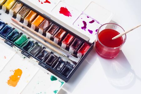 Watercolor paints from natural ingredients in a plastic palette, two brushes and water in a glass cup for washing brushes are on a white table.の写真素材