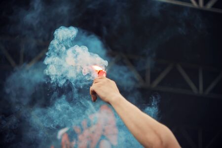 At a rock concert near the stage, a human holds a red signal fire in his hand, which emits thick...