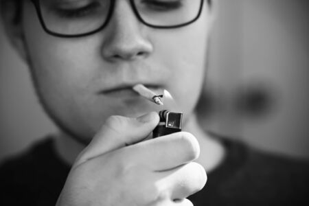 A black-and-white image of a young sad guy lighting a cigarette in his mouth with a black gas lighter.の写真素材