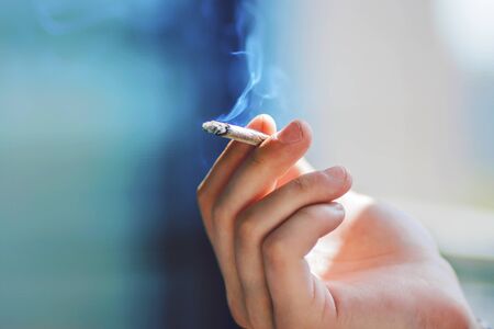 The man's hand holds a cigarette that emits blue harmful tar and nicotine smoke, which is illuminated by bright sunlight.の写真素材