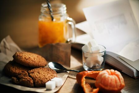 On the wooden table the morning Breakfast - warm sea buckthorn morse in a glass mug, oatmeal cookies, half-peeled tangerine and lump sugar in a Cup, and next to it lies an open book with poems.の写真素材