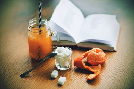 On the wooden table there are pieces of sugar in a Cup, a tangerine in the skin, a glass mug with sea buckthorn sweet morse and a book of poems.の写真素材