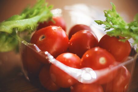 In a small plastic container are ripe cherry tomatoes, and next to them green lettuce leaves, illuminated by bright sunlight.の写真素材