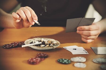 A man is sitting at a table holding cards in one hand and flicking the ash from his cigarette into a...