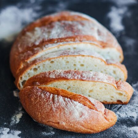  Delicious homemade wheat bread, cut into pieces, lies on a black tray strewn with flour. Home...
