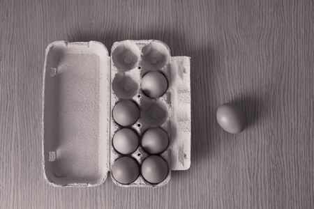 A black-and-white image of chicken eggs lying in a cardboard box, which stands on a wooden table,...