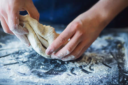 A man in a blue apron is kneading dough with his hands on a dark baking tray sprinkled with flour.