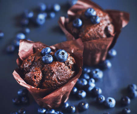 Delicious chocolate cupcakes with blueberry filling stands on a dark blue background among ripe sweet blueberries. Sweet pastries.の写真素材