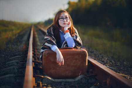 A beautiful young woman with glasses of the Mongoloid race is sitting on the rails with an old suitcase and waiting for the train to arrive. The long wait for the journey. The train left.の写真素材