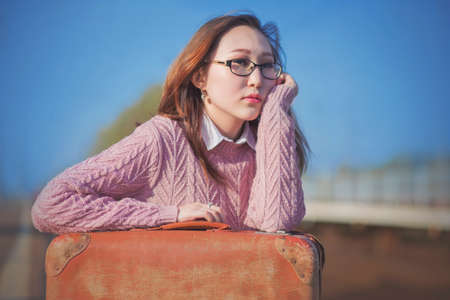 A beautiful elegant young woman in glasses and a pink wool sweater sits with an orange retro suitcase at the station and waits for the train to arrive on a sunny day. Departure.の写真素材