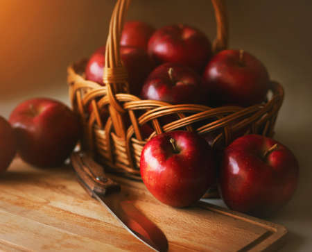 On the kitchen table is a wicker basket with ripe red sweet apples, and next to it is a wooden board and a knife that will cut apples. Still-life. Holiday.の写真素材