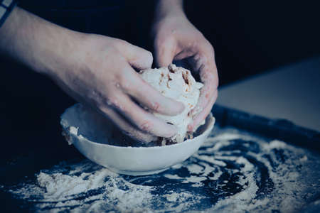 A male chef in a blue apron kneads dough with flour in a white bowl. Making bread dough. The process of home cooking and baking.の写真素材