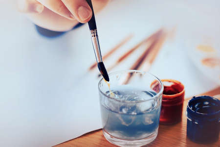 The artist washes the brush in a glass of water, washing off the blue gouache from it. Drawing on a white sheet of paper. Creativity and art. Time for a hobby.の写真素材