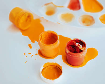 On a white table there is a white palette stained with paint, and next to it there are jars of gouache in red and yellow, one of which is spilled. Creative mess. Drawing and art.の写真素材