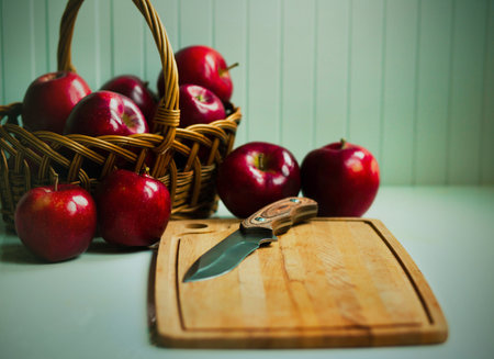 On a white table there is a beautiful wicker basket with scarlet delicious ripe apples, and next to it is a wooden cutting board and a sharp knife. Fruit harvest. Household items in the kitchen.の写真素材