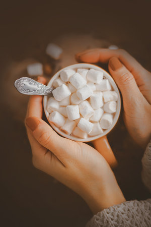 A woman with a brown manicure in a white wool sweater is holding a beige mug with warm cocoa and sweet marshmallow. A pleasant delicious warming drink in the cold season. Hygge.の写真素材