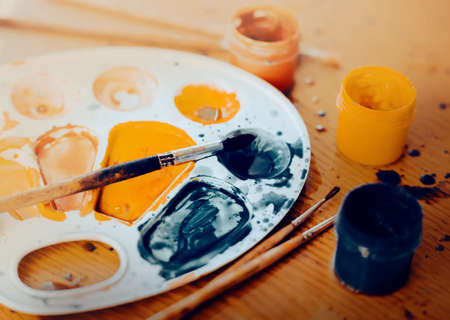 There is a creative mess on the wooden table. The palette is stained with gouache paints, paintbrushes are lying next to them and jars with paints are standing. Creativity and inspiration.の写真素材