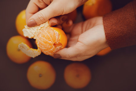 A woman in a brown woolen jacket holds a ripe delicious tangerine in her hands and peels it from the peel. Fruit for Christmas in the cold winter. New Year.の写真素材