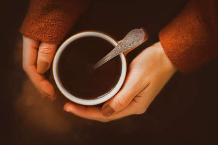 A woman with a brown manicure and a brown woolen warm jacket is holding a beige mug with hot invigorating coffee and a silver patterned spoon. A pleasant start to the day with a strong coffee drink.の写真素材