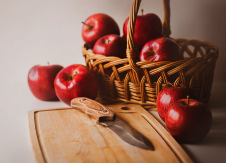 A wicker wooden basket with ripe scarlet delicious apples stands on a white table next to a wooden cutting board on which a sharp knife lies. Proper nutrition. fruit harvest. Vegetarian diet.の写真素材
