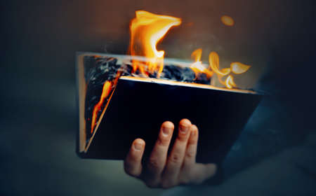 The man holds in his hand a black open book, the pages of which burn with a bright flame and smoke. The Antichrist burns a religious book. censorship.の写真素材