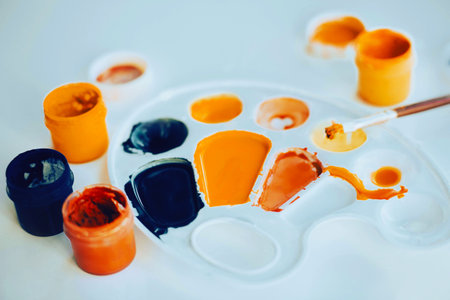 On a white table there is a white palette stained with paint, and next to it there are jars of gouache of different colors and a paint brush. creative mess. The process of drawing.の写真素材