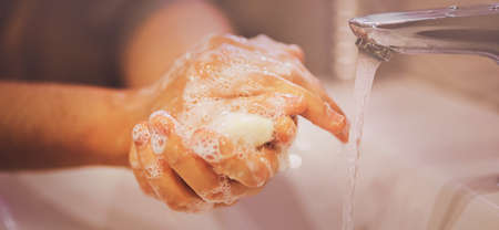 A man holds a white soap in his hands and carefully washes his hands with it over a white sink into which water flows from an open tap. Cleanliness and hygiene.の写真素材