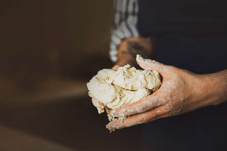 A man in a blue apron holds fresh wheat dough in his hands to make a delicious pizza out of it. home cooking. The baking process.の写真素材