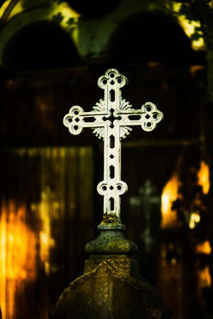 A white old Christian cross stands on a stone pedestal in the shade of tree leaves in the cemetery. Religion and death. Christianity.の写真素材