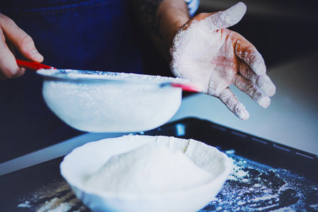 A man in a blue apron sifts flour through a sieve into a white plate. The process of cooking at home. Flour and pastries.の写真素材