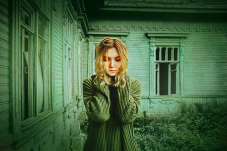 A sad woman in a green sweater stands against the background of an abandoned forsaken wooden green house overgrown with grass. Loneliness against the background of abandoned housing.の写真素材