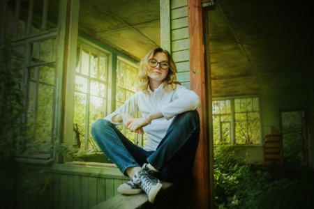 A cute blonde girl in blue jeans and sneakers is sitting on the wooden railing of an old wooden...