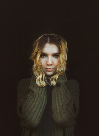 A beautiful blonde woman in a vintage wool sweater stands on a black background and looks with an evil mysterious look. Mysticism and horror.の写真素材
