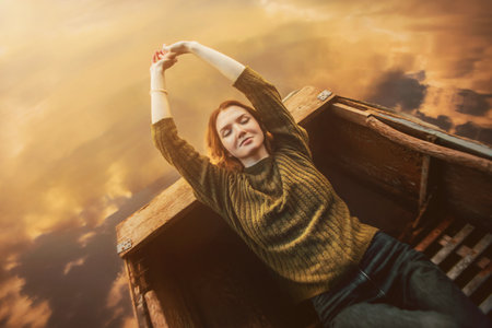 A beautiful blonde woman in a vintage sweater, with her eyes closed, lies in an old rowing boat that floats on the water surface of a fabulous lake, in which the sky and clouds are reflected at dawn.の写真素材