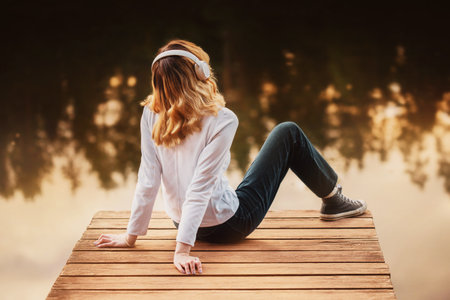 A beautiful blonde woman in a turtleneck and jeans is sitting on a wooden pier with headphones and listening to music, relaxing near the water on a sunny evening. Pleasure and tranquility. Harmony.の写真素材