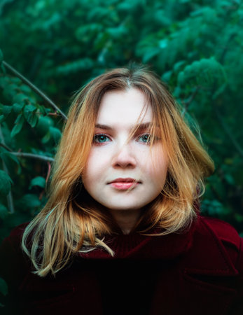 A close-up portrait of a beautiful young blonde woman in a burgundy coat against the background of green foliage of trees. lifestyleの写真素材